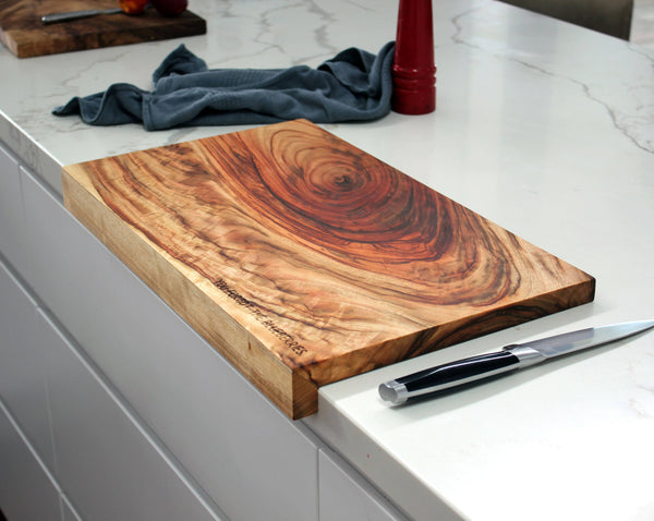 Wooden cutting board on a kitchen counter with a knife and towel in the background